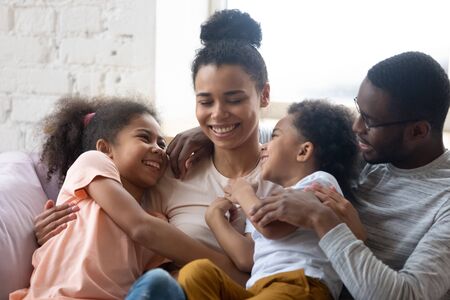 African american happy family with two kids enjoy time together. Cute diverse daughter and son with parents sit on warm comfy couch in living room. Young father and mother have fun with adorable kidsの写真素材