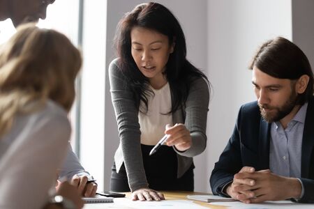 Confident Asian female team leader talk explain business idea to diverse colleagues at meeting, motivated young ethnic businesswoman speak negotiate brainstorm with multiracial coworkers at briefingの写真素材