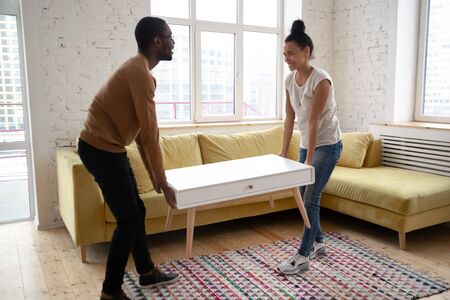 Young happy african american couple moving wooden table. Attractive family of diverse woman and man relocation furniture in new flat. Husband and wife perform permutation. First time buyersの写真素材