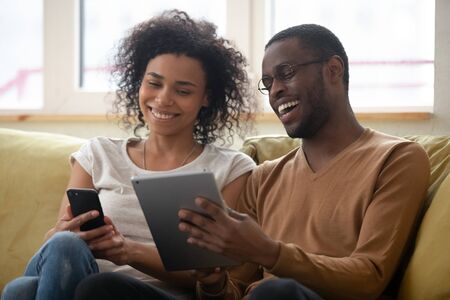 Laughing millennial african american man and woman couple looking at smartphone screen. Smiling and happy husband and wife watching funny online video together in social network.の写真素材