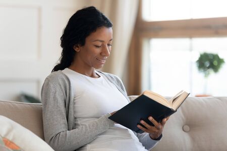 Calm and smiling, young african american girl read interesting book, sit on comfortable sofa in living room, enjoy relaxed weekend, spend free time at home, study literature, leisure and hobbyの写真素材