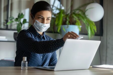 Young female employee wearing protective face mask disinfect computer with sanitizer, woman cleanse sanitize laptop with antibacterial liquid, protect from COVID-19, coronavirus pandemic conceptの写真素材