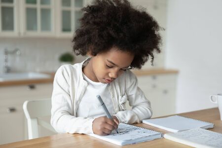 Small African American girl sit at desk in kitchen handwrite in notebook having distant class, smart little biracial kid prepare homework study alone on quarantine at home, homeschooling conceptの写真素材