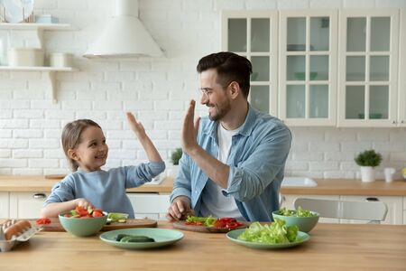 Excited young Caucasian father and little daughter give high five engaged in funny activity cooking salad at home together, happy dad and small girl child have fun preparing healthy food in kitchenの写真素材