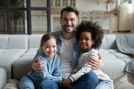 Portrait of young caring father sit on couch hug cuddle with small multiracial daughters, happy Caucasian dad embrace play with multiethnic little children, enjoy family weekend at home togetherの写真素材