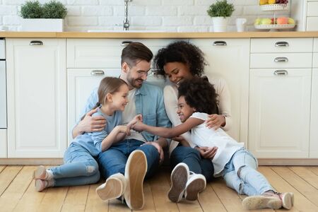 Happy multiracial young parents relax on warm wooden floor in kitchen with little daughters, smiling family with small girls children sit rest in modern design renovated home, have fun togetherの写真素材