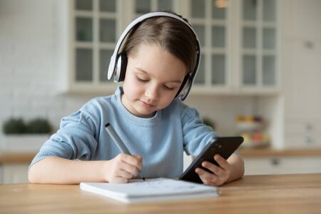 Smart little Caucasian girl in headphones do homework online class on smartphone, small child in...