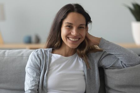 Head shot portrait close up beautiful young woman with healthy toothy smile sitting on cozy sofa at home, pretty girl looking at camera, touching hair, posing on comfortable couch in living roomの写真素材