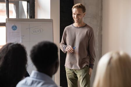 Smiling Caucasian male trainer make flipchart presentation interact with diverse colleagues at...