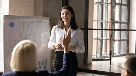 Smiling millennial businesswoman interact with diverse employees making flip chart presentation in office, young female coach or trainer ask questions at briefing, cooperation, interaction conceptの写真素材