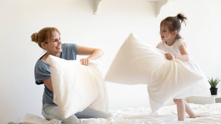 Happy young mother pillows fight and laughing with daughter on bed at bedroom. Smiling family of mom and little cute girl having fun and playing with cushions at home together.の写真素材