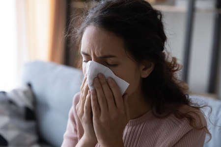 Close up head shot unhealthy young lady using paper tissue, wiping runny nose. Unhappy stressed woman suffering from seasonal allergy or caught cold, blowing nose or coughing in handkerchief.の写真素材