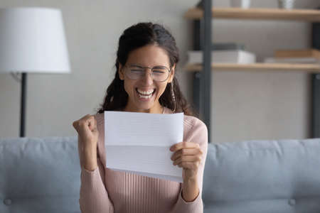 Overjoyed young attractive lady in eyewear holding paper letter, celebrating business success. Emotional millennial woman making yes gesture, feeling excited about personal achievement at home.の写真素材