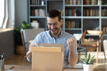 Excited happy man looking into cardboard box, sitting at work desk, received parcel with awaited online store order, smiling laughing young male celebrating success, lottery winner prizeの写真素材