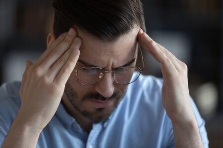 Close up unhealthy stressed young man wearing glasses touching forehead, suffering from headache, tired overworked employee, student massaging temples, feeling pain, health problemの写真素材