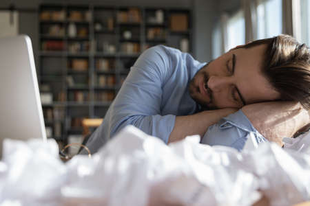 Close up tired exhausted man sleeping at workplace, lying on hand with closed eyes, sitting at desk with laptop and crumpled papers, working on difficult project, unproductive young male dozingの写真素材