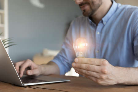 Close up confident businessman holding light bulb in hand, sitting at wooden desk with laptop,...