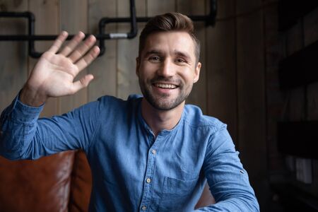 Headshot portrait of happy Caucasian young man sit in cafe wave greeting talking on video call,...