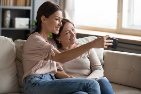 Smiling middle aged mother and daughter taking selfie together, hugging and sitting on couch at home, young woman and happy older mature mum photographing, two generations good relationsの写真素材