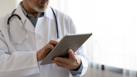 Old male doctor holding modern digital tablet computer standing in hospital, close up view. Senior middle aged professional medic wears white coat and stethoscope using healthcare ehealth application.の写真素材