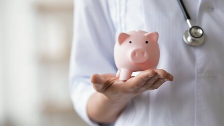Woman doctor wearing white coat and stethoscope holding piggy bank box in hands. Healthcare services expenses, money savings on health insurance, medicine cost concept. Close up view, copy spaceの写真素材