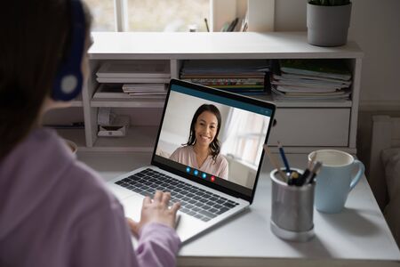 Back view of young girl sit at desk at home talk have online video call lesson with teacher or tutor, teenage schoolgirl engaged in webcam conversation, study distant use web conference app on laptopの写真素材