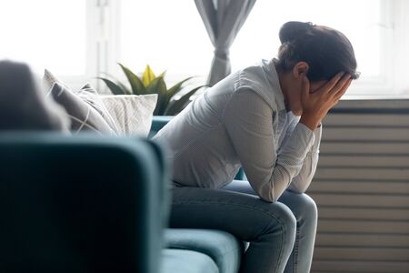 Side view unhappy depressed young indian girl hiding face in hands, sitting on sofa alone at home. Desperate millennial mixed race woman suffering from personal life problems or stress, headache.の写真素材