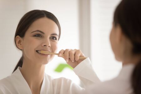 Young Caucasian woman look in mirror in bathroom brush teeth with toothbrush toothpaste, millennial...
