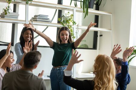 Happy attractive businesswoman conduct training with diverse colleagues. Diverse team celebrating business success at meeting. Smiling female mentor with raised hands congratulating colleagues.の写真素材