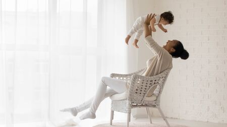 Happy loving african American young mother sit in chair at home relax play with funny little newborn baby girl, smiling biracial mom have fun lift in air enjoy family weekend with cute infant childの写真素材