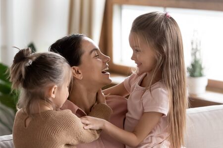 Happy mother having fun with two little daughters at home, sitting on cozy couch in living room, laughing young mum holding cute preschool girls, enjoying weekend with adorable childrenの写真素材
