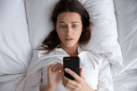 Top view awakened woman lying in bed holds smart phone looks at screen watching time overslept and late feels stressed, upset female read received awful news, being cheated threaten or hacked conceptの写真素材