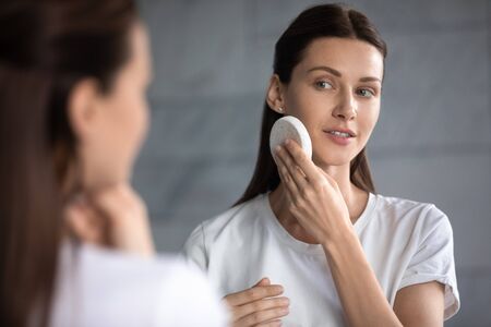 Woman reflected in mirror view over shoulder, female cleansing purify sensitive skin holds facial sponge, soft hygienic daily usage, dirt make up effective removal, massage procedure, skincare conceptの写真素材