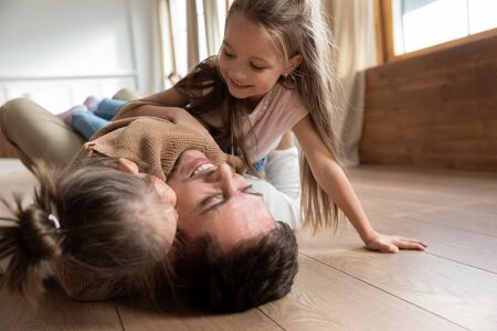 Happy father lying on warm floor with two adorable little daughters close up, hugging and cuddling, family having fun at home, spending free time together, cute preschool girls playing with dadの写真素材