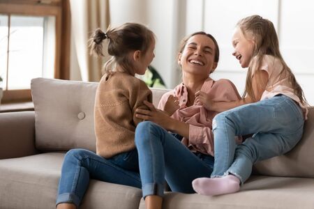 Happy mother playing with two little daughters, sitting on couch at home, cute preschool girls...