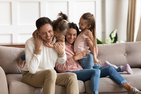 Happy mother and father hugging and cuddling with two little daughters, sitting on cozy sofa at home, smiling parents and preschool children having fun together, family enjoying free timeの写真素材