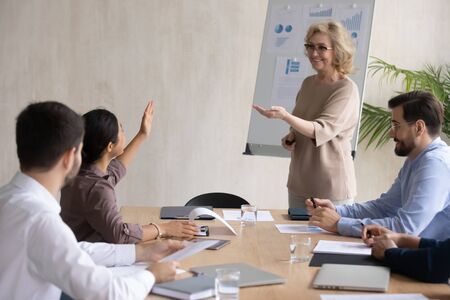 Smiling indian businesswoman raising hand asking mature mentor in boardroom at company meeting....