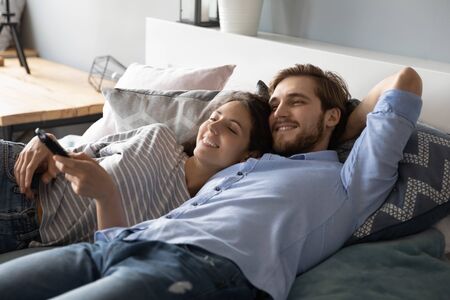 Happy young Caucasian man and woman lying relaxing on comfortable bed at home watching TV together, smiling millennial couple rest in bedroom, enjoy lazy leisure weekend indoors, relaxation conceptの写真素材