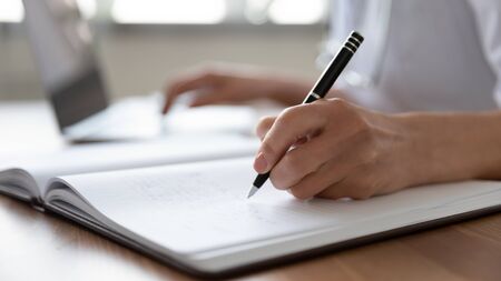 Female professional doctor hand making notes in medical journal using laptop computer sitting at desk. Woman physician, nurse or pharmacist wearing white coat writing in paper notebook. Close up viewの写真素材