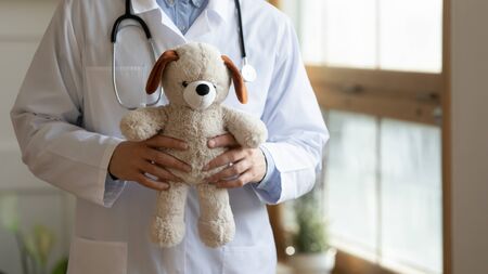 Close up of male child doctor in white medical uniform hold plush teddy bear, man pediatrician stand with fluffy toy, show care and comfort in hospital or private clinic, children healthcare conceptの写真素材