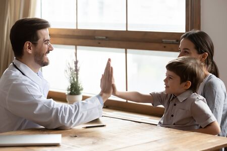 Positive male doctor have fun give high five play with small boy patient visiting with mom, excited little child greeting with caring man pediatrician at consultation, children healthcare conceptの写真素材