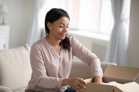 African woman sit on couch prepare parcel for sending to relatives or friends, girl collect personal...