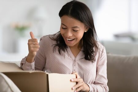 African woman sit on sofa at home opens carton box parcel feels satisfied by delivered goods show...