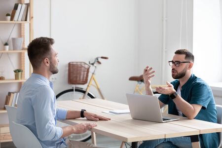 Serious bearded businessman talking and interviews new male applicant using resume and laptop. Hr...