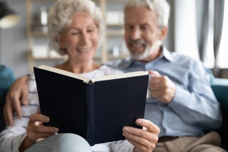 Focus on paper book bestseller in retired family hands. Happy middle aged married couple reading novel together, cuddling relaxing on couch, enjoying retirement weekend free leisure time at home.の写真素材