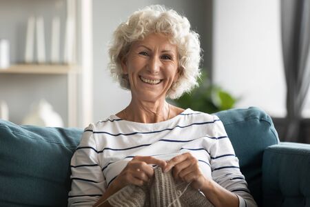 Happy elderly mature beautiful hoary grandmother relaxing on comfortable sofa, knitting warm sweater with woolen threads. Smiling middle aged woman enjoying free hobby weekend time alone at home.の写真素材