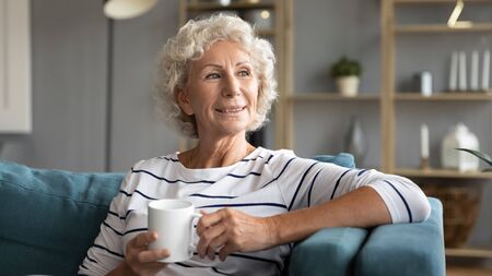 Head shot smiling old senior woman relaxing on comfortable couch with mug of hot tea, enjoying free...
