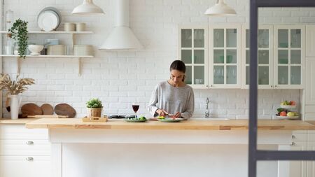 Young Caucasian woman stand at modern design kitchen counter cooking vegetable salad drinking wine, millennial girl female tenant or renter prepare vegetarian food in beautiful renovated homeの写真素材