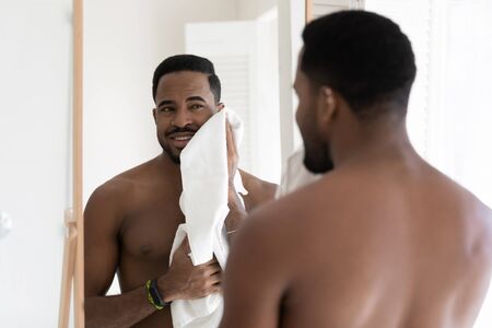 Naked young african American man look in mirror in bathroom rub wipe body with towel after shower, handsome metrosexual biracial male do morning daily beauty procedures in bath, hygiene conceptの写真素材