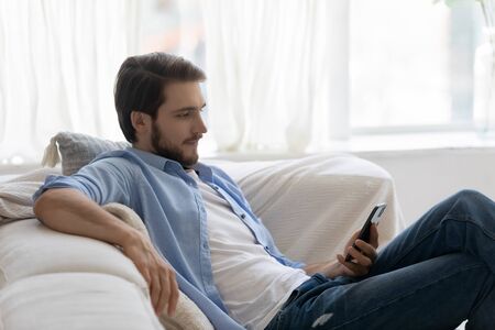 Young Caucasian man sit relax on sofa at home use modern smartphone gadget, relaxed millennial male rest on comfortable couch in living room browse wireless internet on cellphone, technology conceptの写真素材